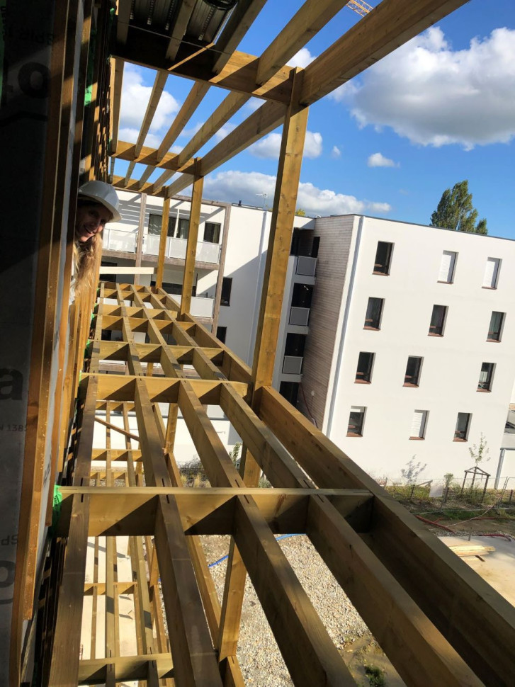 Photo prise par la fenêtre, on voit le balcon : poutres en bois, pas encore de "sol". La tête d'un coopérateur dépasse par la fenêtre. Ciel bleu. Immeuble voisin en fond